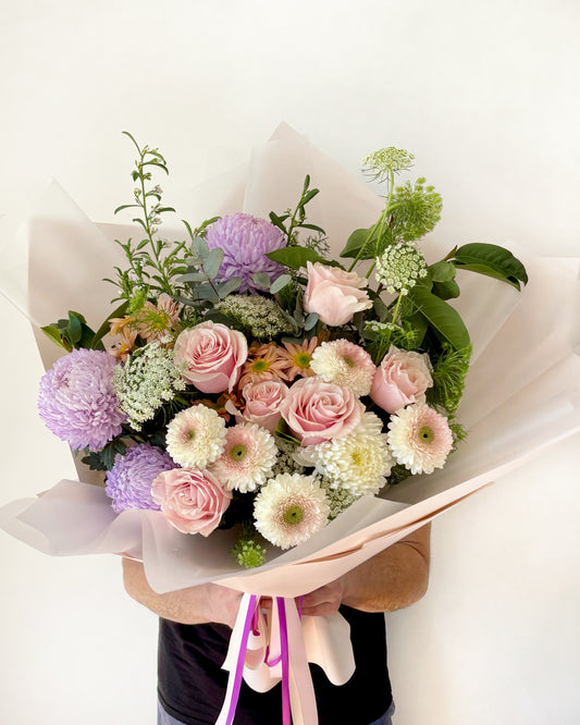 A bouquet of flowers including roses and other seasonal flowers in pastel colors like pink, purple, and white, wrapped in a neutral-colored paper with a pink ribbon.