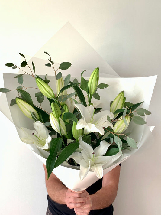 A bouquet of Oriental lilies with green leaves, wrapped in white paper, with a focus on the stunning blooms.